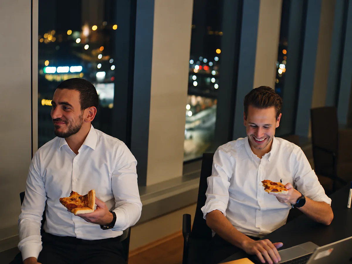 To menn i hvite skjorter spiser pizza på kontoret en kveld, med byens lys bak