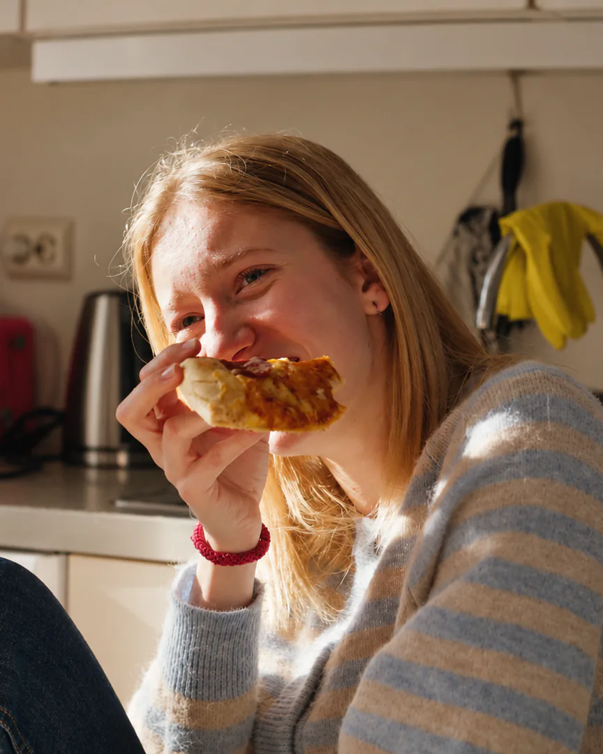 En jente ler og tar en bit av et pizzastykke på kjøkkenet