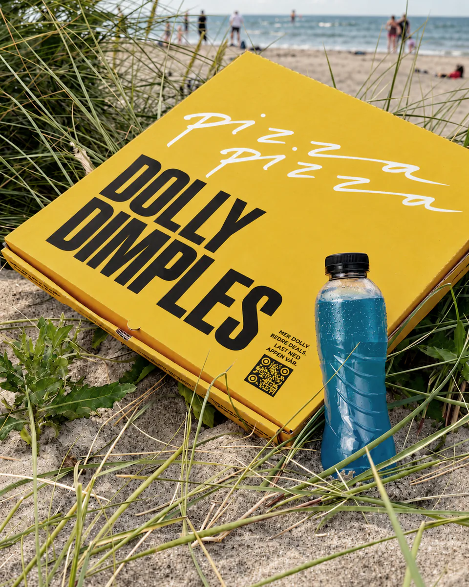 En gul Dolly Dimples-pizzaeske i sanddynene på en strand, med en flaske sportsdrikk