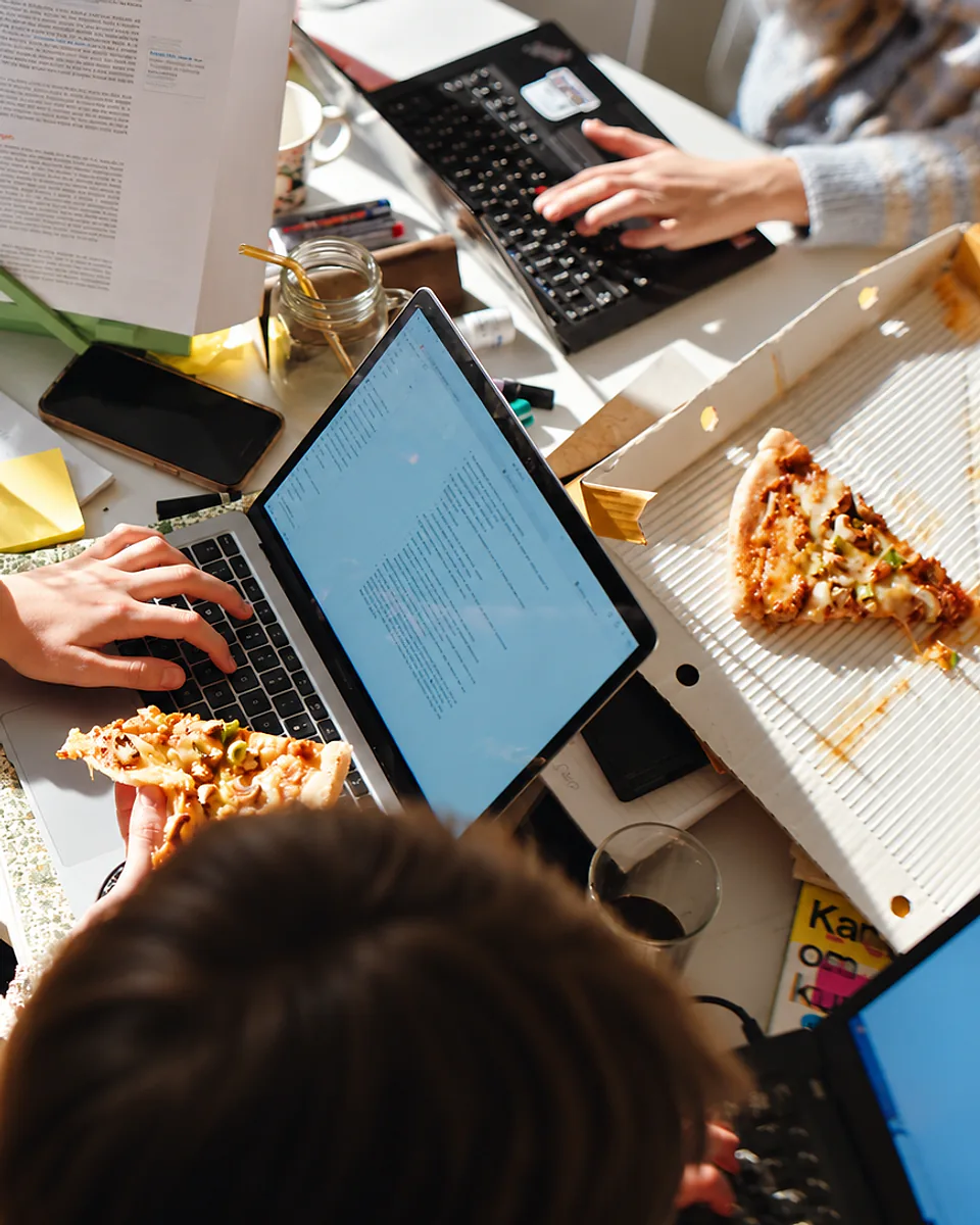 Kolleger med bærbare PC-er og pizza rundt et bord fullt av papirer