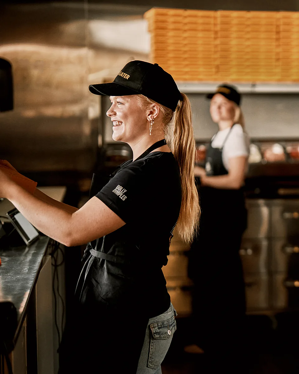 En blid ansatt tar imot en bestilling, mens kollegene lager pizza på kjøkkenet bak